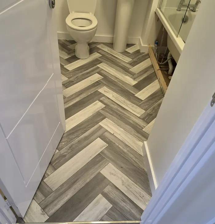 Grey and light wood-effect herringbone-pattern flooring in a bathroom, with a threshold strip at the doorway