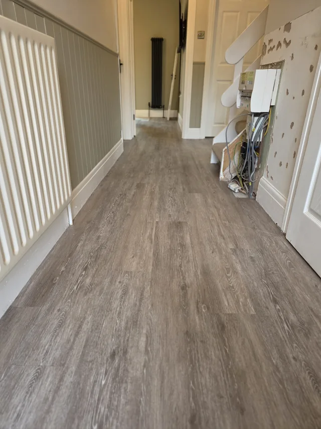 Wood-effect full-plank LVT along the hallway beside a white radiator, wall panelling and the stairs