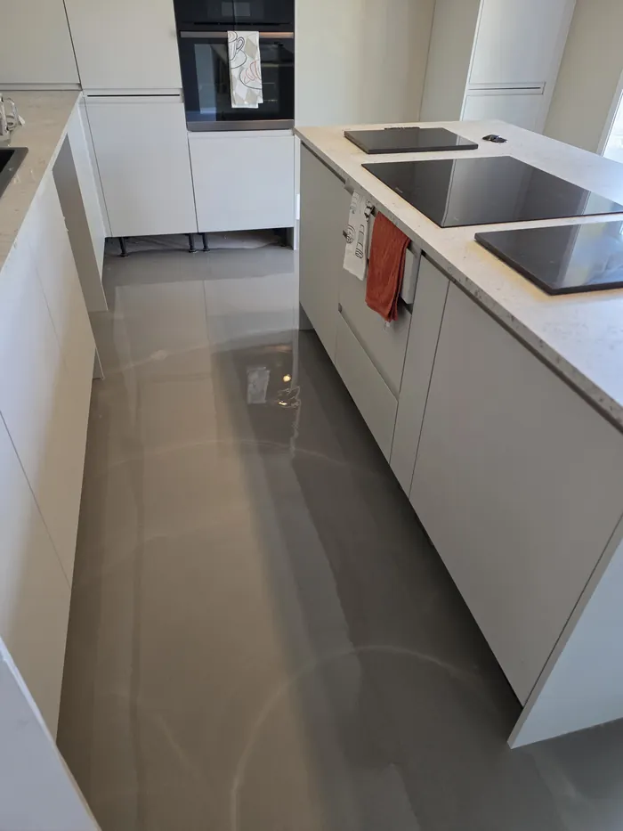 Fresh grey levelling compound before LVT fitting between pale kitchen cabinets and an island, with reflections across the floor