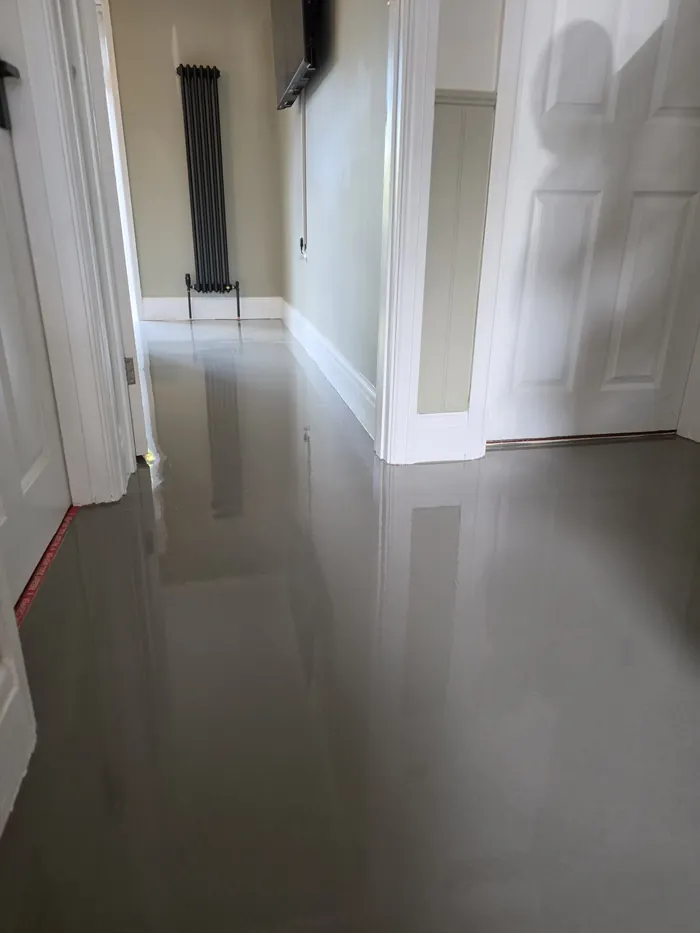 Fresh grey levelling compound before LVT fitting in a hallway facing a black vertical radiator beside a white door