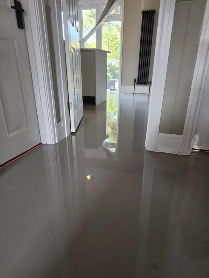 Fresh grey levelling compound before LVT fitting, viewed through a hallway doorway towards a bright window and black vertical radiator