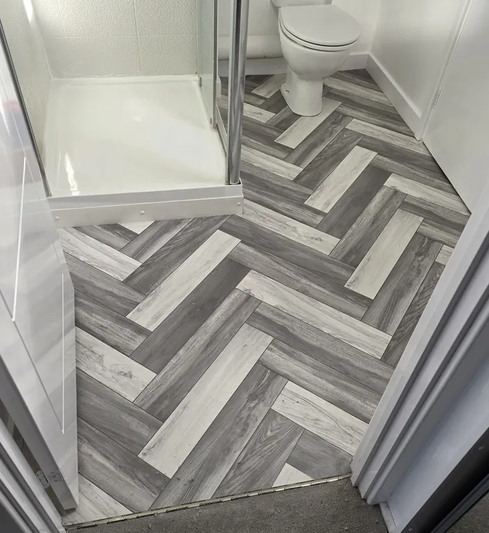 Grey and light wood-effect herringbone-pattern flooring laid through a small shower room, seen from the doorway